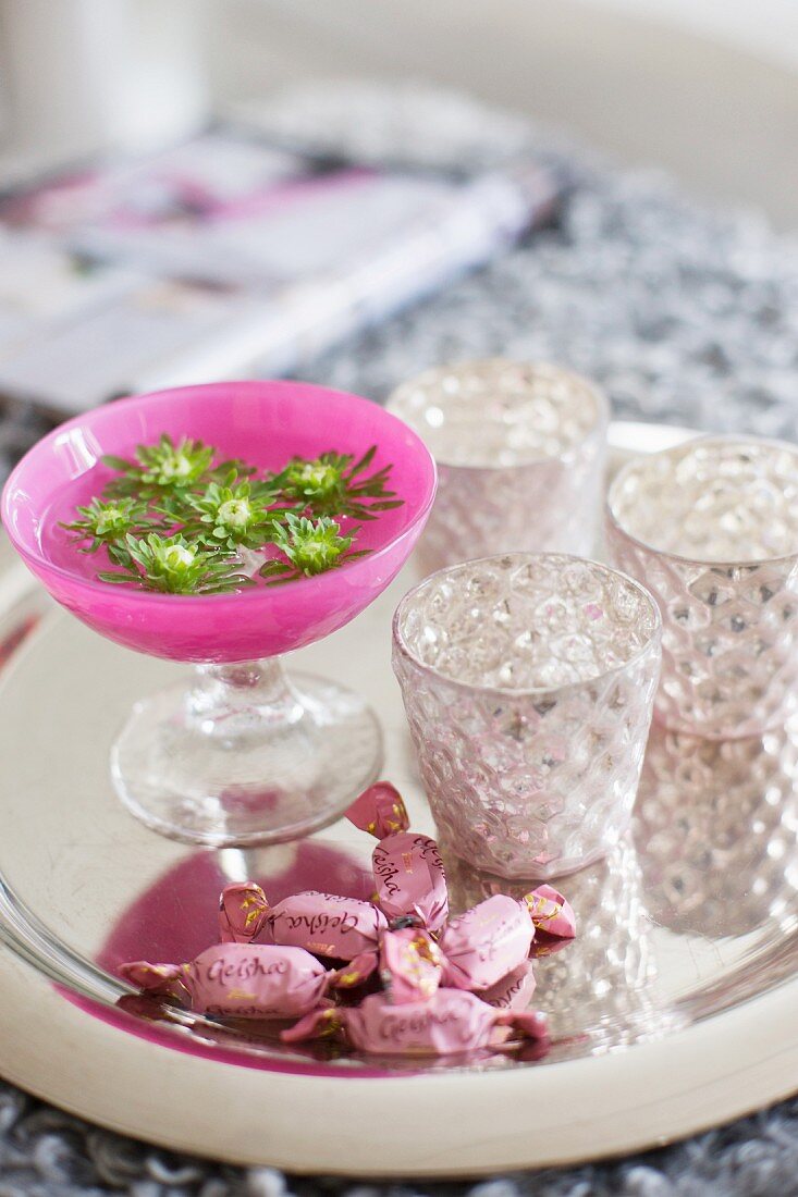 Pinkfarbener Kelch mit Blüten, silbrig schimmernde Teelichter und Bonbons auf einem glänzenden Tablett