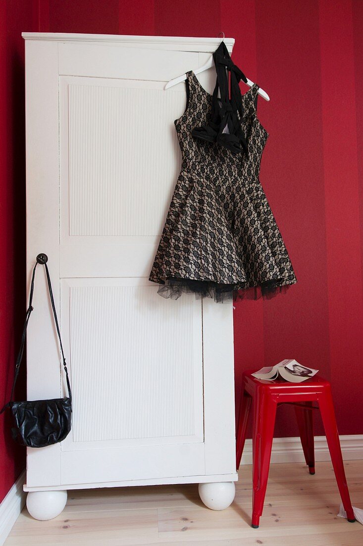 Short cocktail dress and handbag hanging from white wardrobe next to plastic stool against red striped wallpaper