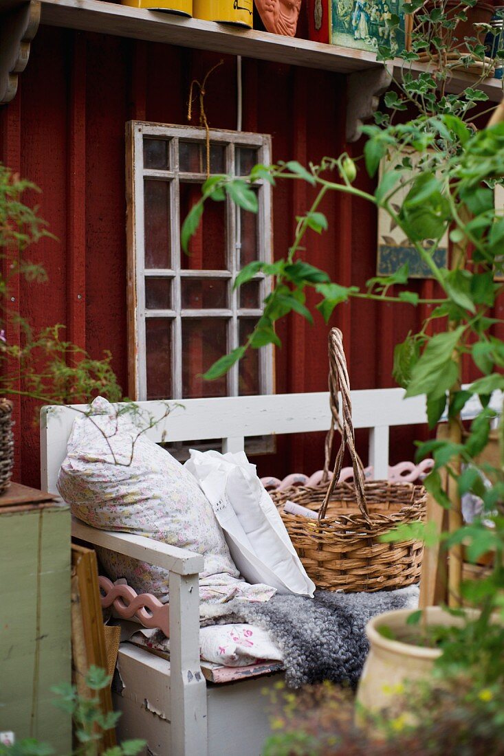 Hellgrau lackierte Holzbank mit Kissen und Korb, im Hintergrund schlichtes Holzhaus
