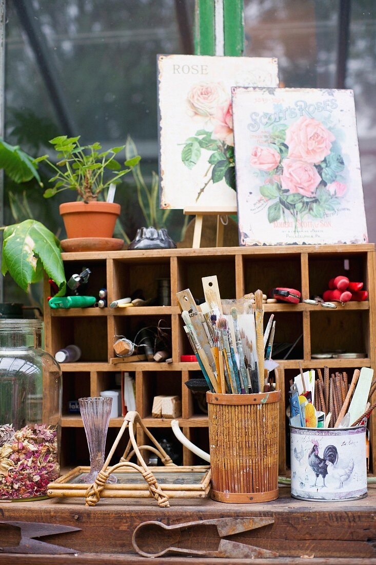Arbeitsplatz mit Behältern vor Regal mit Gartengeräten, obenauf Vintage Schilder mit Rosenmotiv