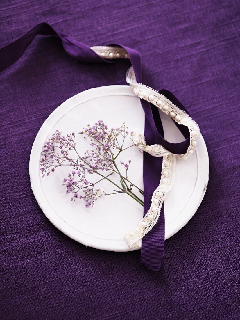 Weisser Teller auf violetter Tischdecke mit violetten Blüten, Satin- und Spitzenband