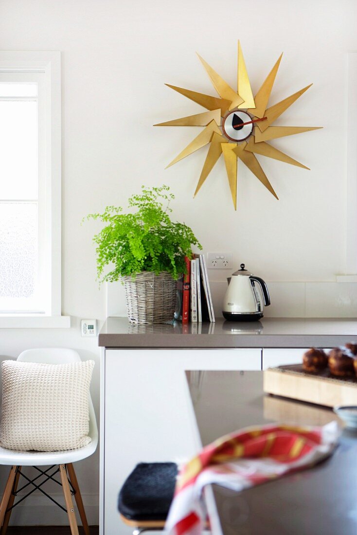View across counter of kitchen unit below star-shaped, brass wall clock and classic shell chair below window
