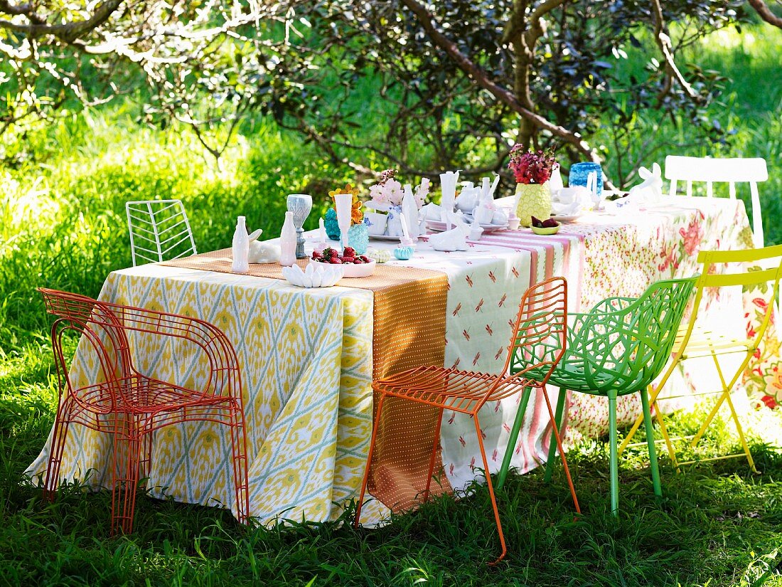 Gedeckter Tisch mit verschiedenen Tischdecken und farbigen Metallstühlen auf Wiese im Garten