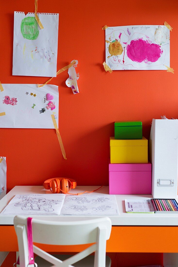Aufbewahrungsschachteln aus farbigem Karton auf Schreibtisch, vor orangeroter Wand mit Kinderzeichnungen