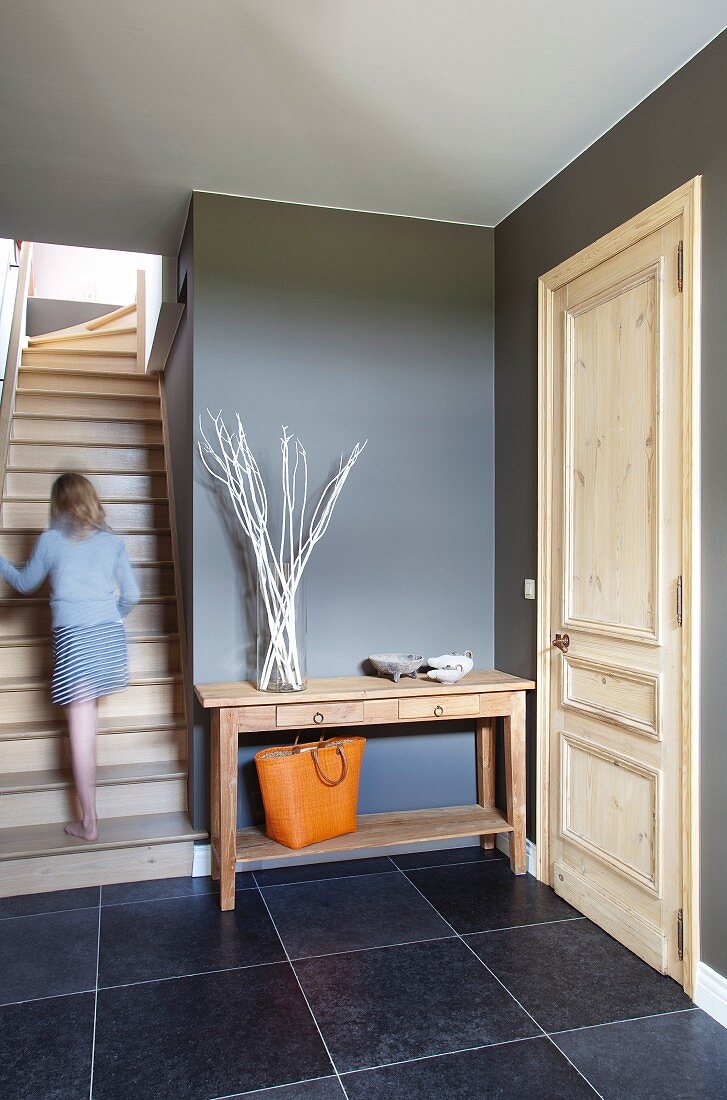 Foyer with pale wooden door in charcoal grey wall and dark floor tiles; girl walking up stairs