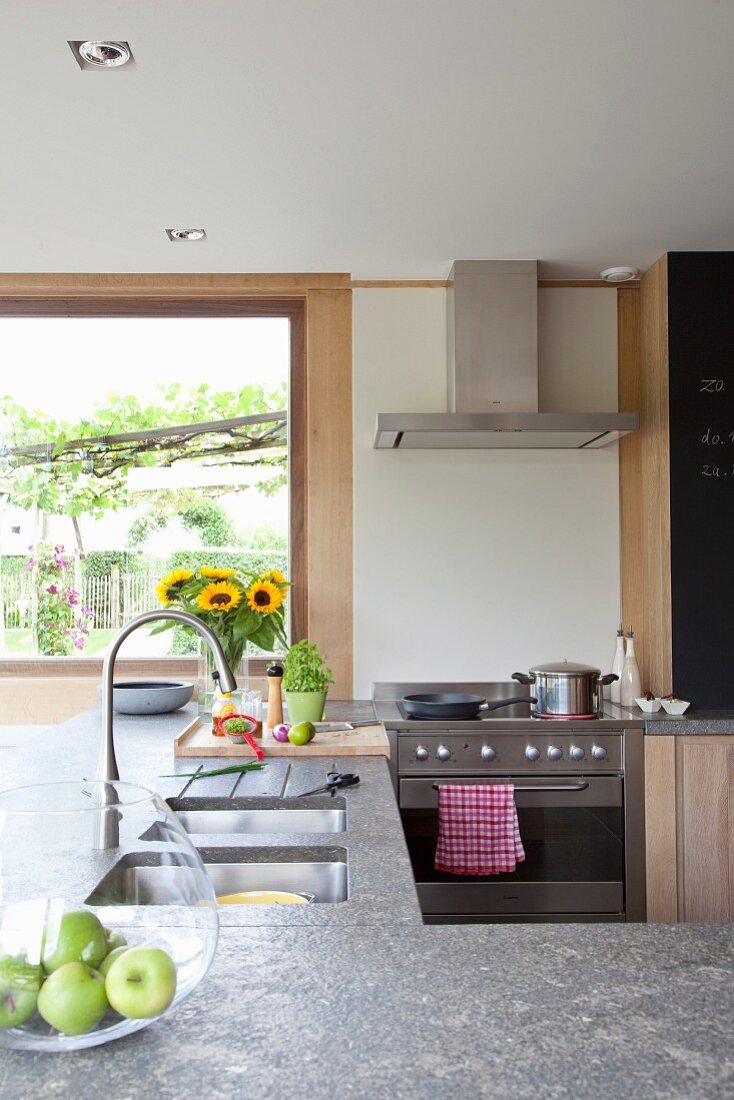 Offene Küche mit Arbeitsplatte aus Naturstein und Spülbecken, grosses Fenster zum Garten im Hintergrund