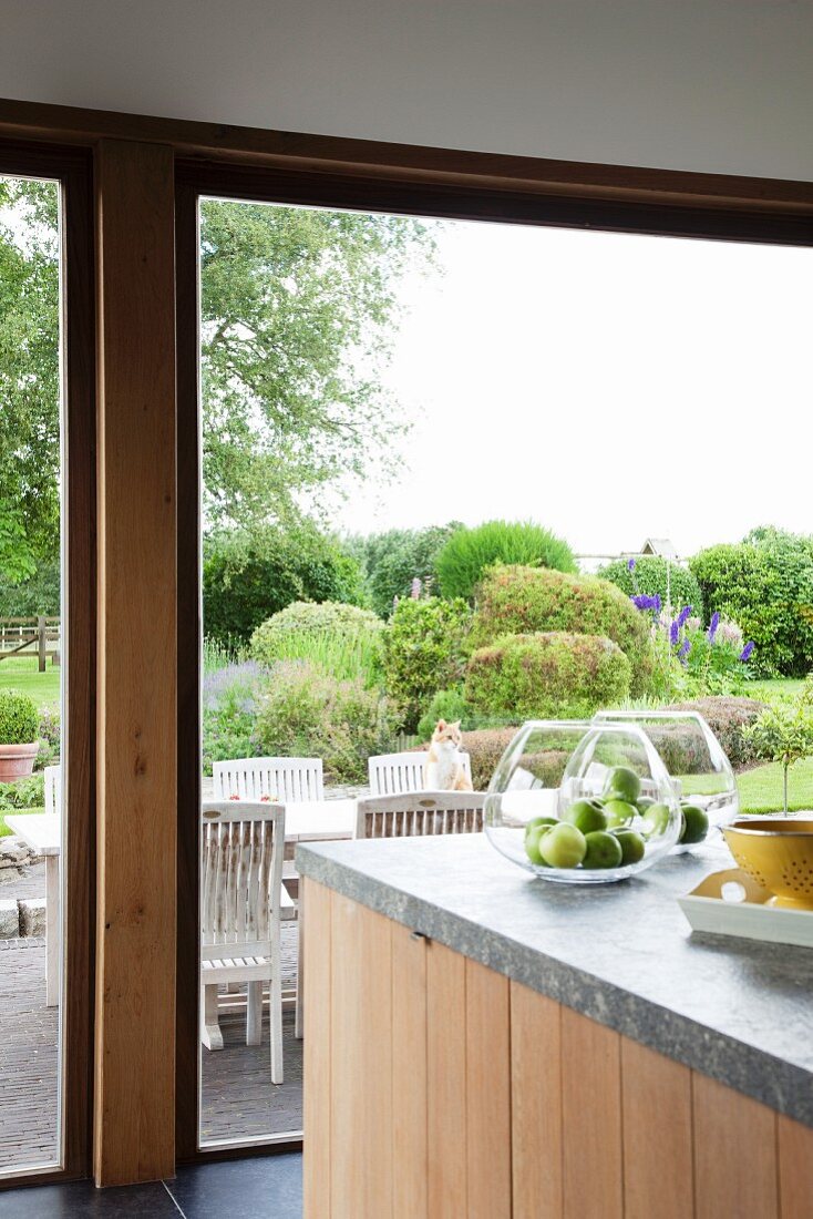 Äpfel in Glasschüsseln auf Natursteinplatte der Kücheninsel, dahinter Fensterfront zur Terrasse und dem großen Garten