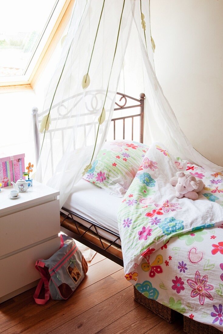 Zarter Vorhang über verspieltem Kinderbett aus Metall und Bezüge mit stilisiertem Blumenmuster