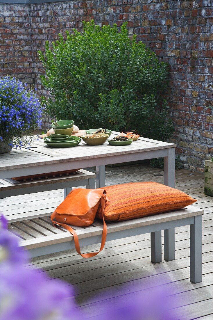 Damentasche und Kissen auf Holzbank vor passendem Tisch auf Holzterrasse
