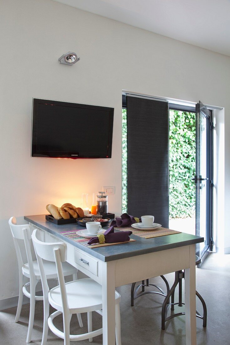 Frühstücksgedecke auf rustikalem Küchentisch, vor Wand mit aufgehängtem Fernseher, daneben offene Terrassentür