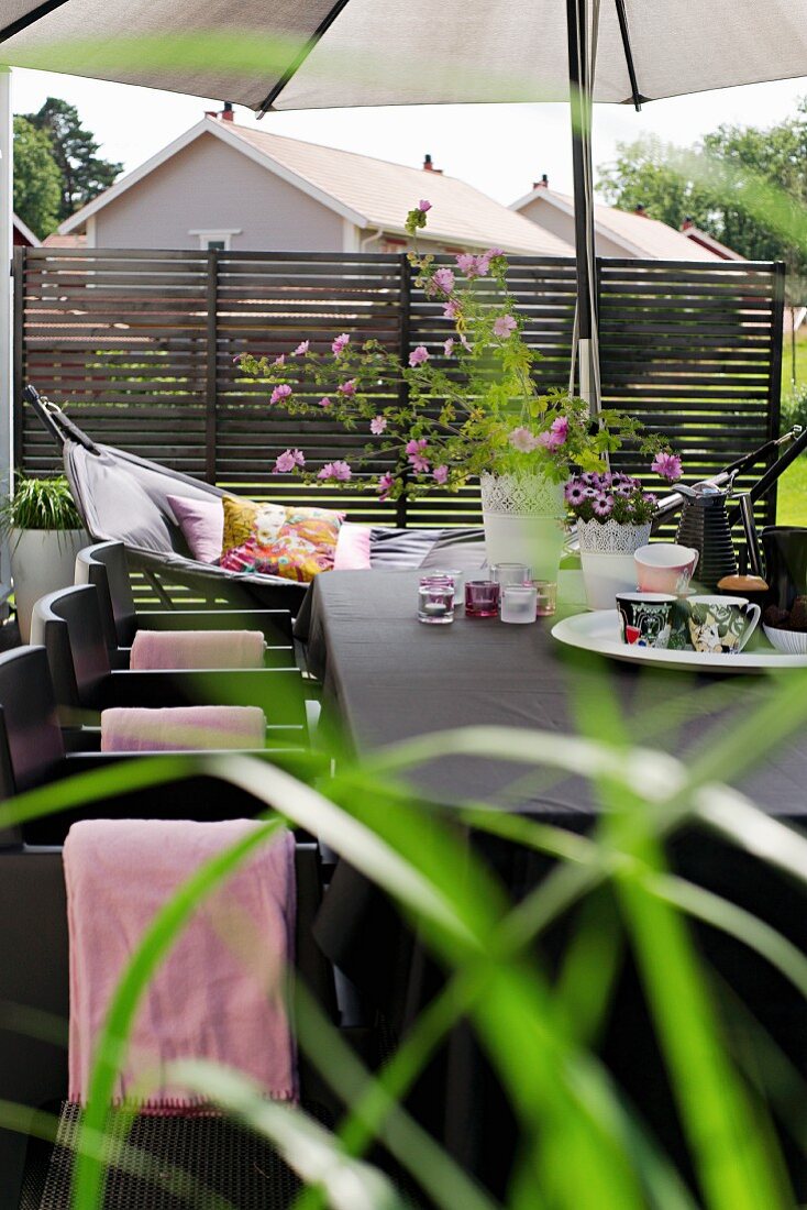 Tisch mit Tassen, Windlichtern und Gartenstrauss unter Sonnenschirm auf der Terrasse, im Hintergrund Hängematte