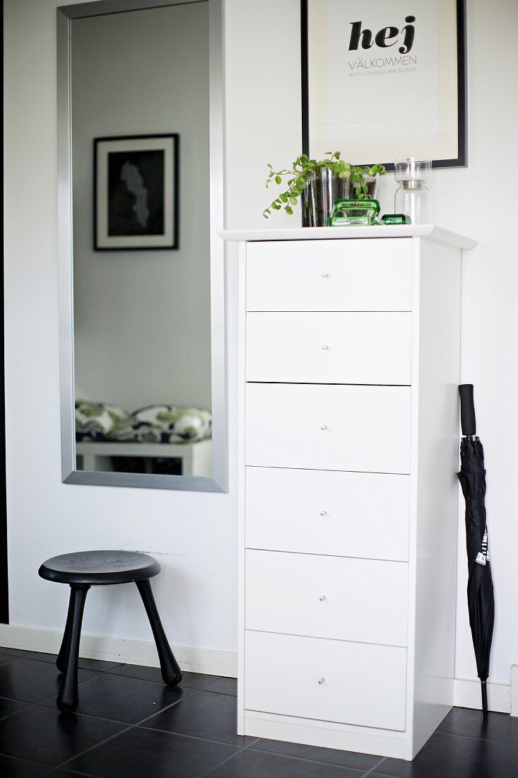 Narrow, white chest of drawers, black umbrella and stool on black tiled floor in hall