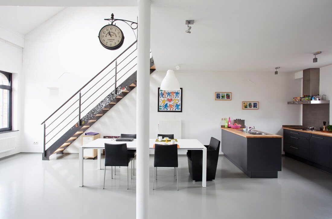 Minimalist, retro loft apartment with white dining table and black chairs in front of staircase and open-plan kitchen with dark grey cabinets