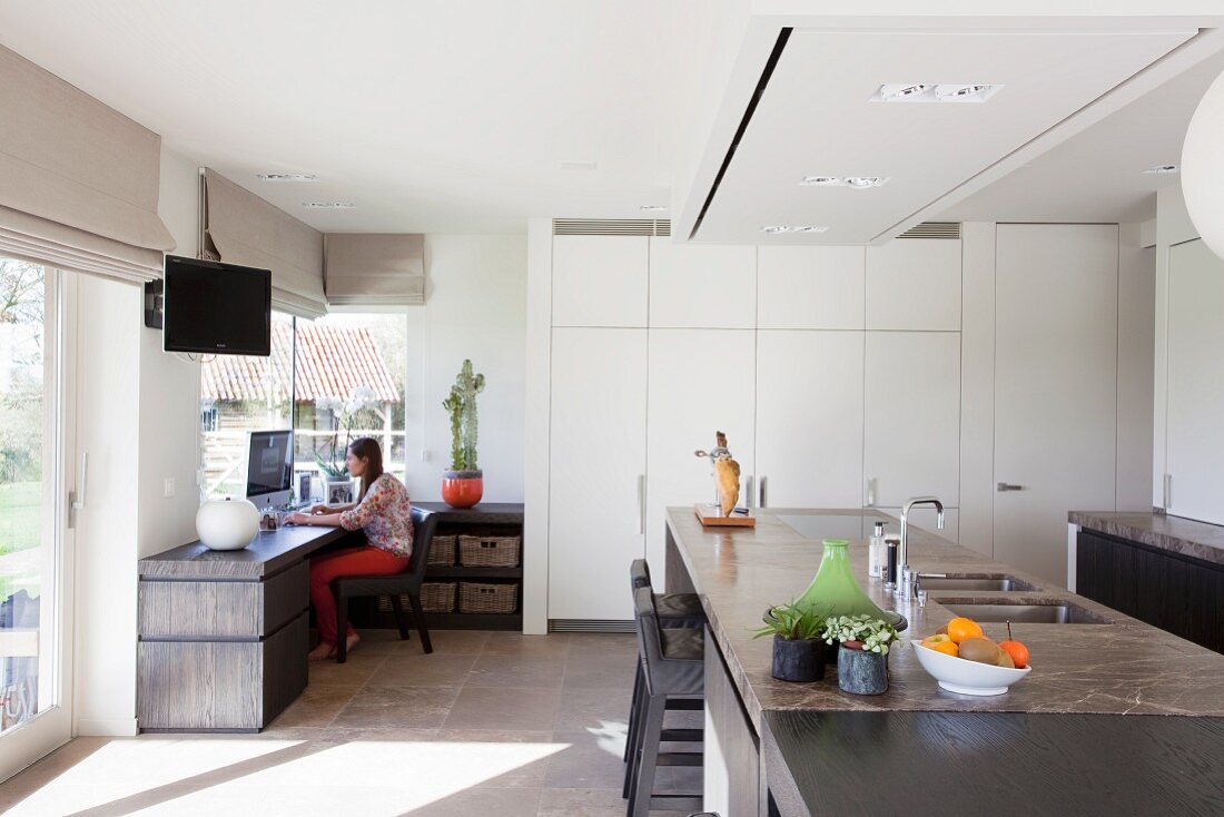 Moderner Wohnraum - Essbereich mit Küchenblock, dunkle Steinplatte, im Hintergrund weisser Einbauschrank, in Zimmerecke Frau am Arbeitstisch vor Fenster