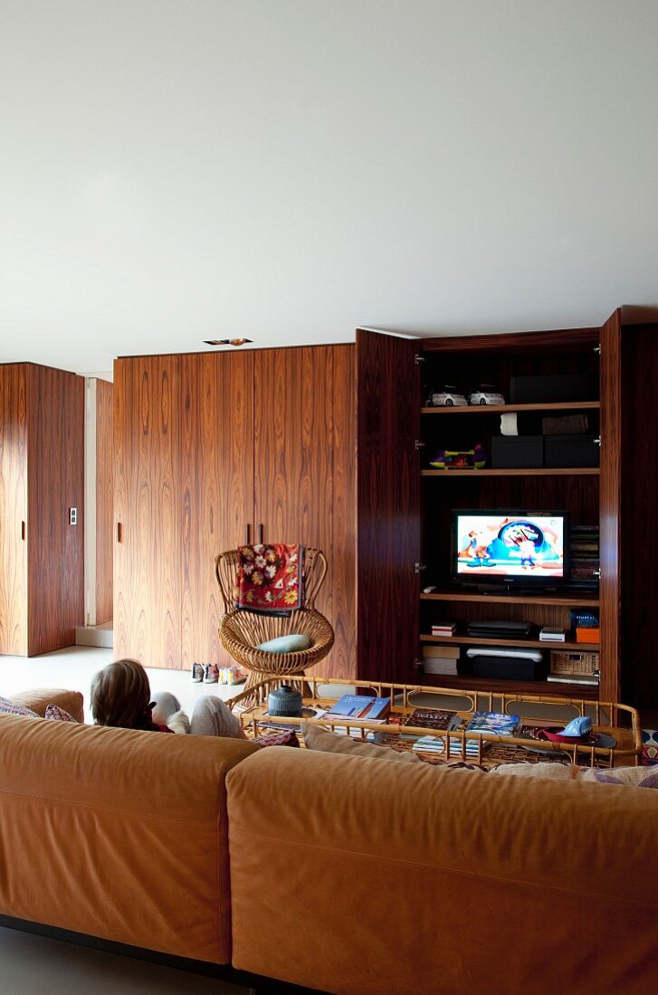 Gemütliche, braune Ledercouch in Loungebereich, Kind beim Fernsehen, Einbauschrank aus Palisanderholz mit Fernseher hinter offener Tür