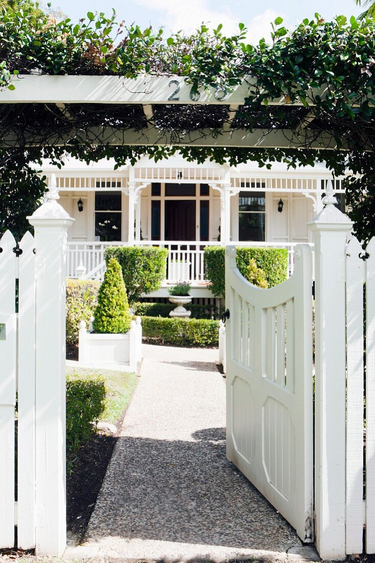 White garden gate below climber-covered pergola and gravel path leading through front garden to Colonial-style country house in sunlight