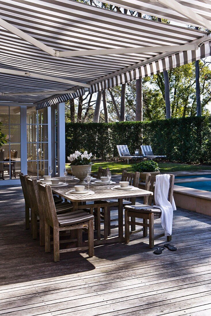 Set table on spacious wooden terrace below striped awning with view of sunny garden