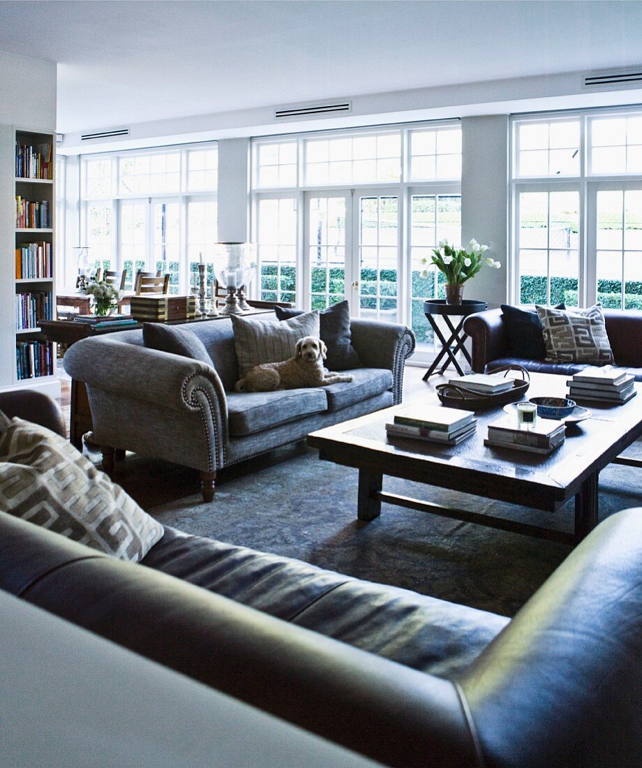Various leather and grey velvet sofas around coffee table in open interior with white French windows