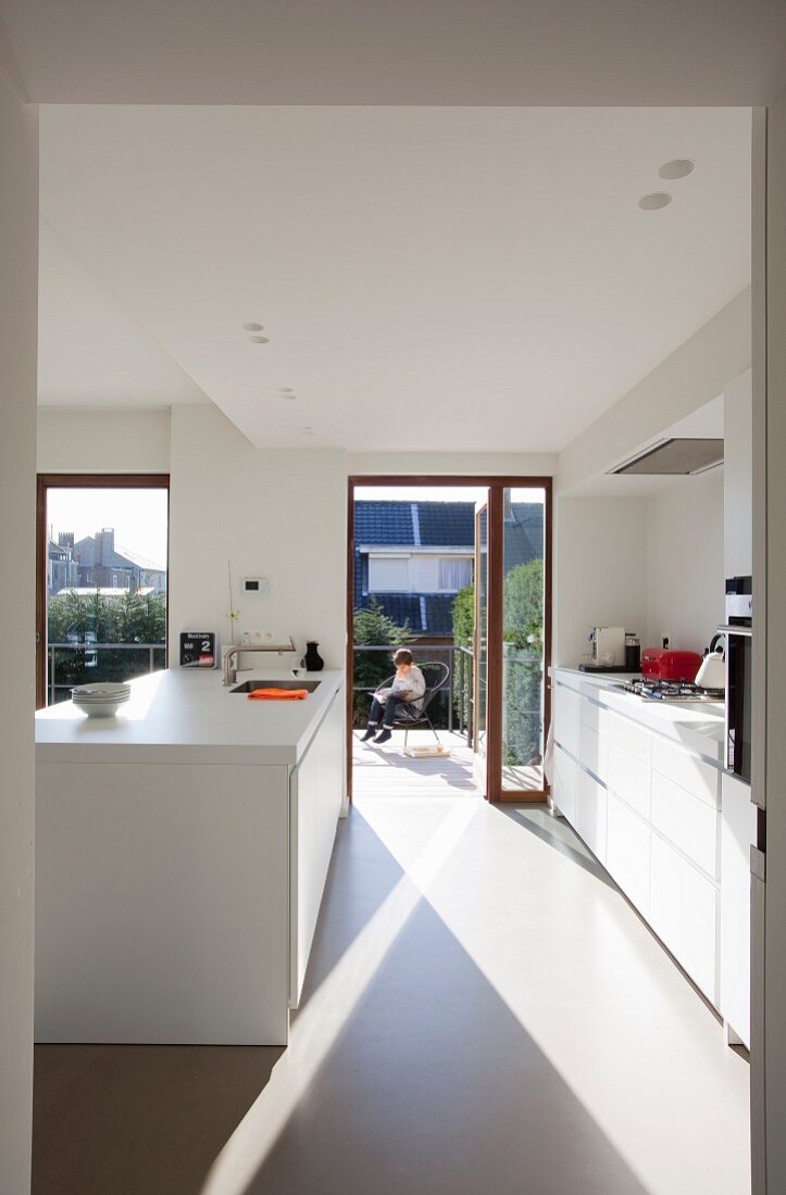 Sonnenbeschienene Designerküche in Weiß, im Hintergrund offene Balkontür und Blick auf kleinen Jungen auf dem Balkon sitzend