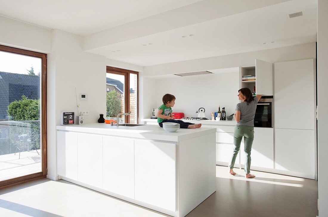 weiße Designer Küche mit minimalistischem Flair, kleines Kind auf Küchenblock sitzend gegenüber Mutter vor Hängeschrank