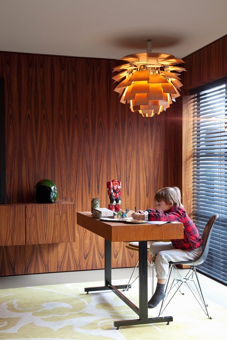 Child sitting at minimalist desk under classic pendant lamp in room with 60s-style wall panelled in rosewood
