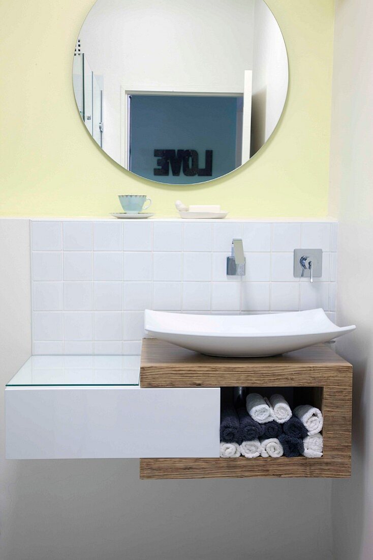Waschschüssel auf Regal-Unterbau aus Holz und weiße Schublade; runder Spiegel an gelb getönter Wand
