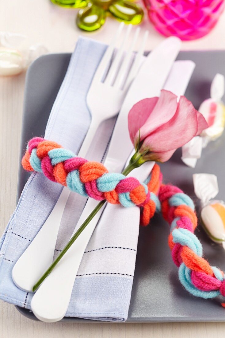 Besteck, Stoffserviette und Blumendeko mit geflochtenem, farbigem Wollstrang als Serviettenring umwickelt