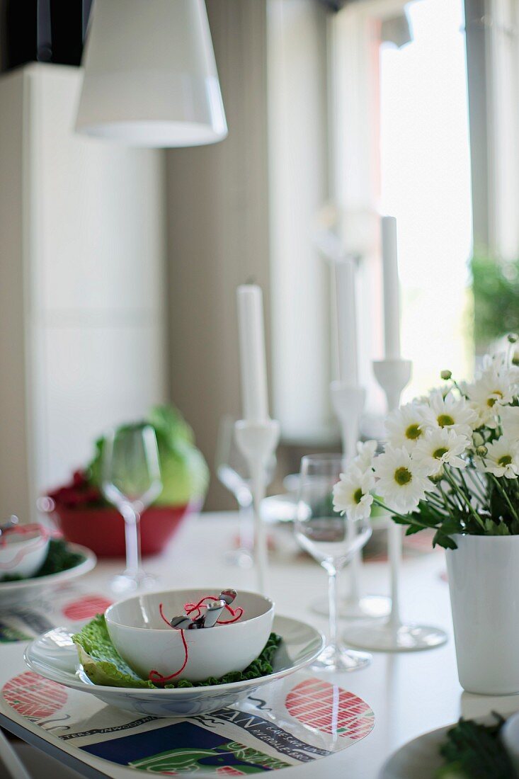 Gedeck mit weisser Schale auf Tischset, seitlich teilweise sichtbarer Blumenstrauss in Vase