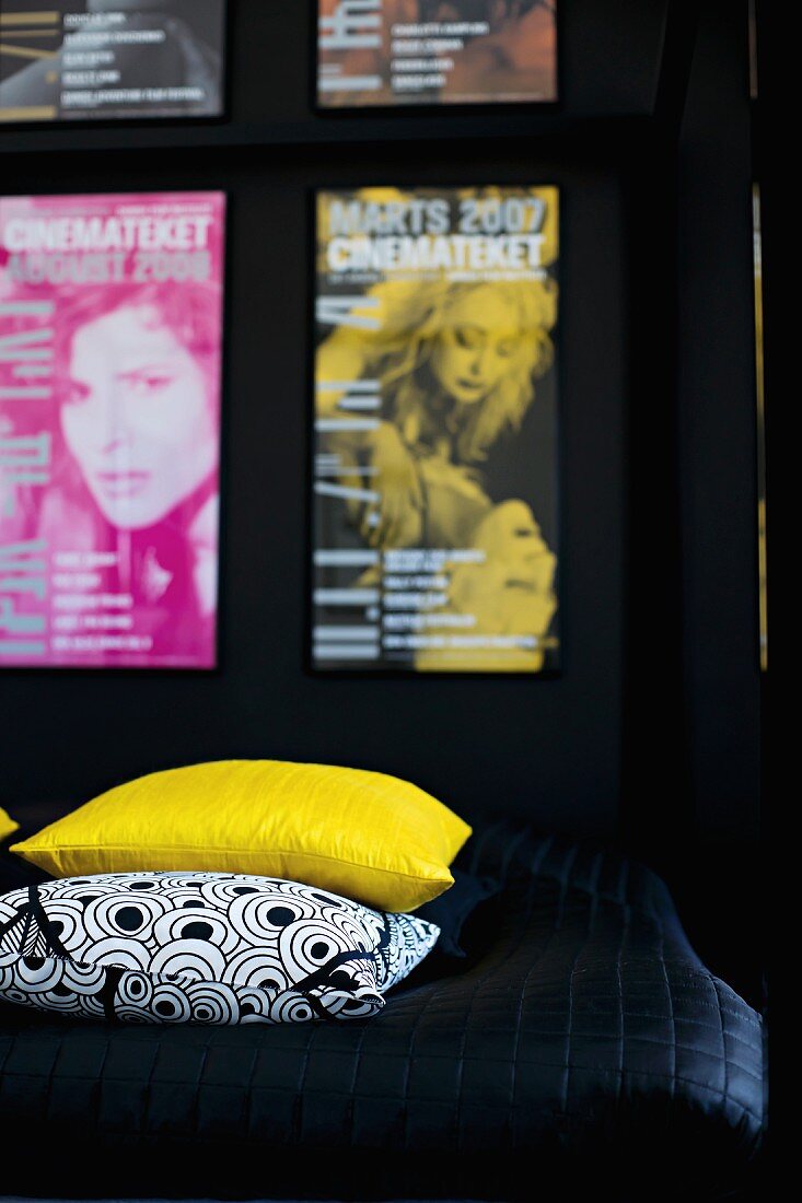 Ausschnitt eines Bettes mit gelbem und schwarzweiss gemustertem Kissen auf schwarzem Plaid, an Wand Poster Sammlung