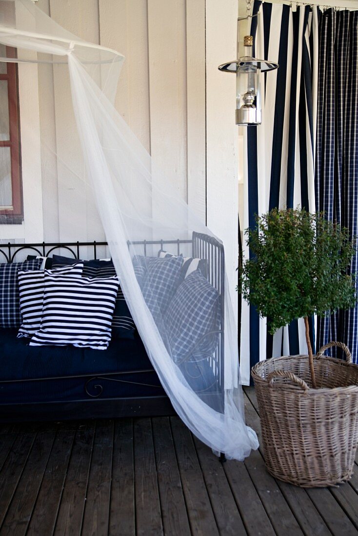 Metall Tagesbett mit weissblauen Kissen und Baldachin, seitlich Bäumchen im Korb auf Holzboden einer Veranda