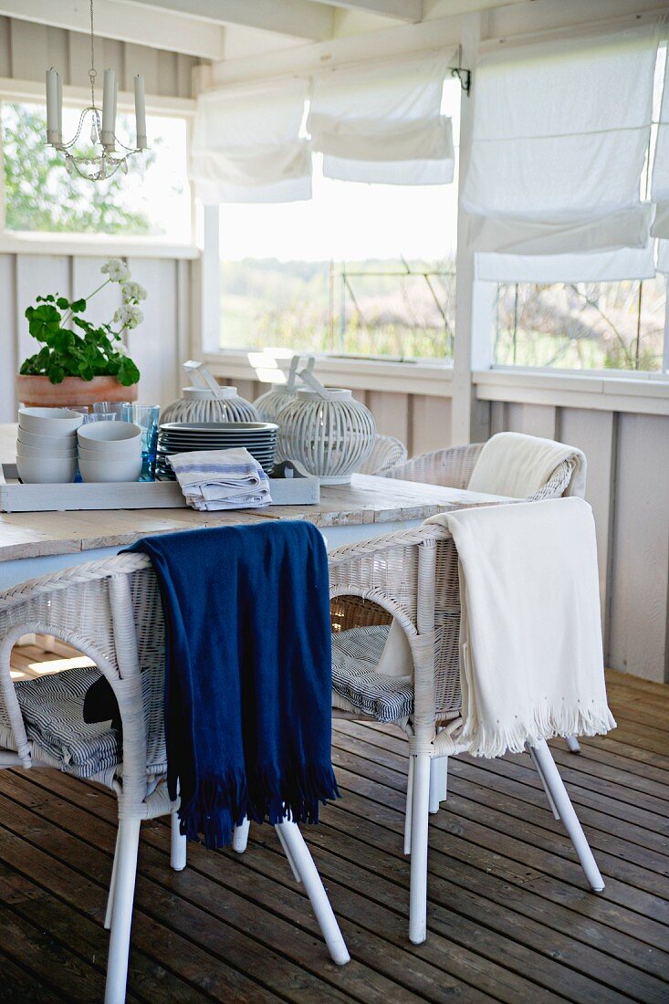 Tablett mit Geschirr auf Tisch, weiße Rattanstühle auf Holzboden einer teilweise holzverschalten Veranda