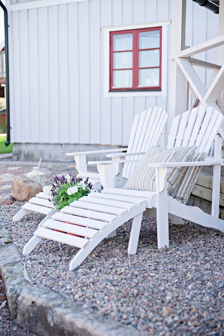 weiße Holz Liegestühle auf gekiestem Terrassenboden, im Hintergrund Holzhaus mit rotem Fenster