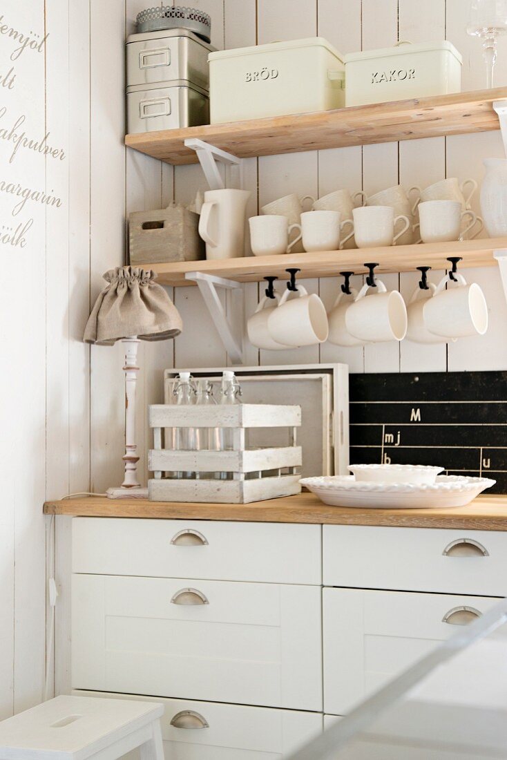 Kitchen counter with white base units below wooden shelves of storage containers and cups hanging from hooks