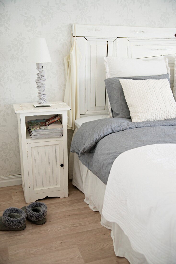 White wooden bedside table next to bed with tall headboard and grey and white bed linen