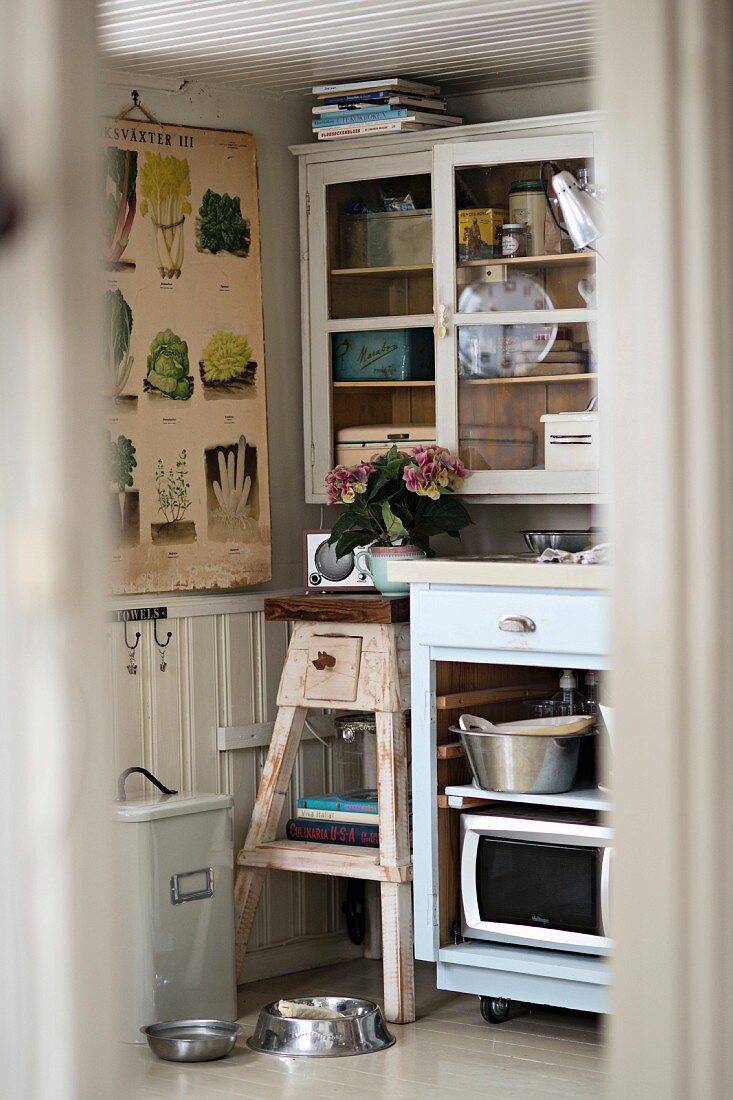 Blick durch Türspalt in Küchenecke, rustikaler Blumenständer neben teilweise sichtbarem Unterschrank, darüber Vitrinen Hängeschrank