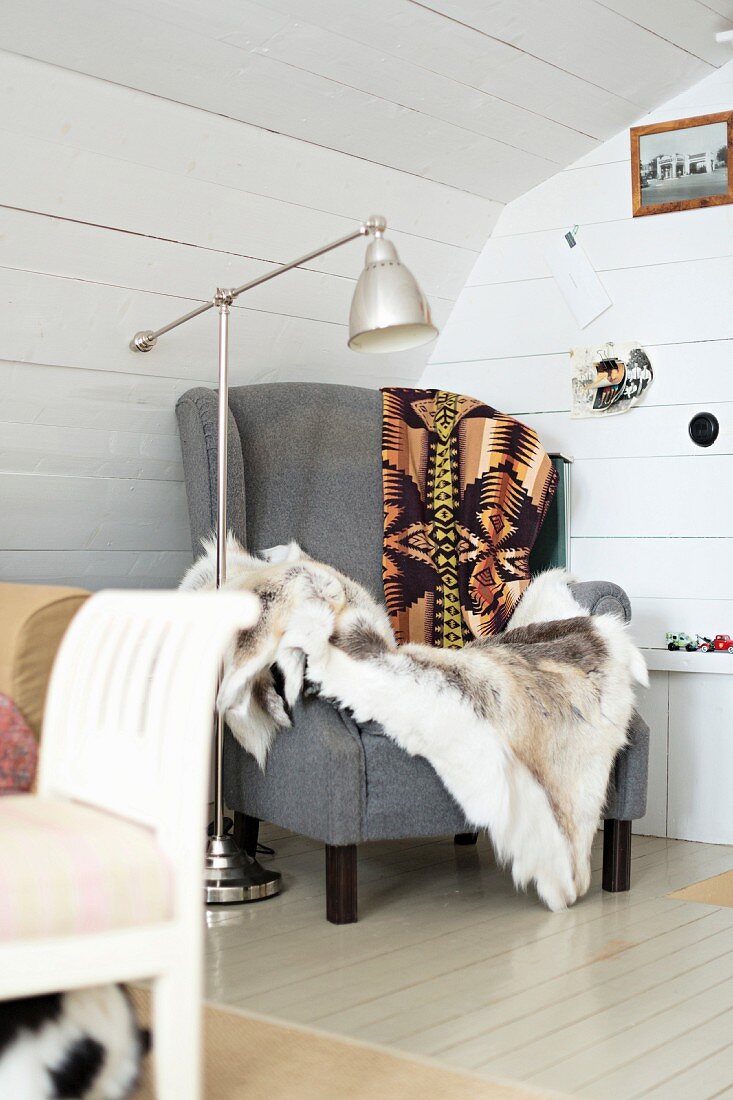 Comfortable grey armchair with fur blanket and retro standard lamp in corner of room with white wooden walls