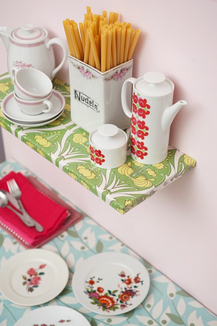 Geschirr und Porzellanbehälter mit Nudeln auf mit Wachstuch bezogenem Regalboden an rosa getönter Wand