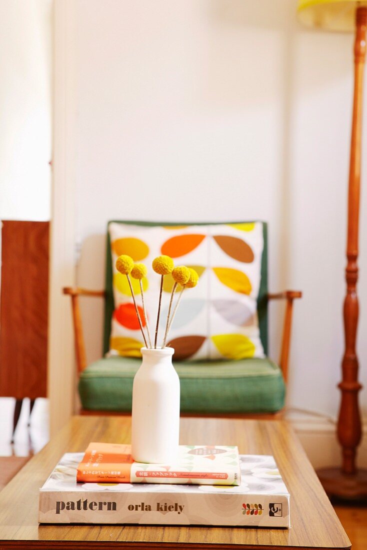 Simple vase of dried yellow flowers on stacked books and coffee table; 50s armchair with patterned scatter cushions in background
