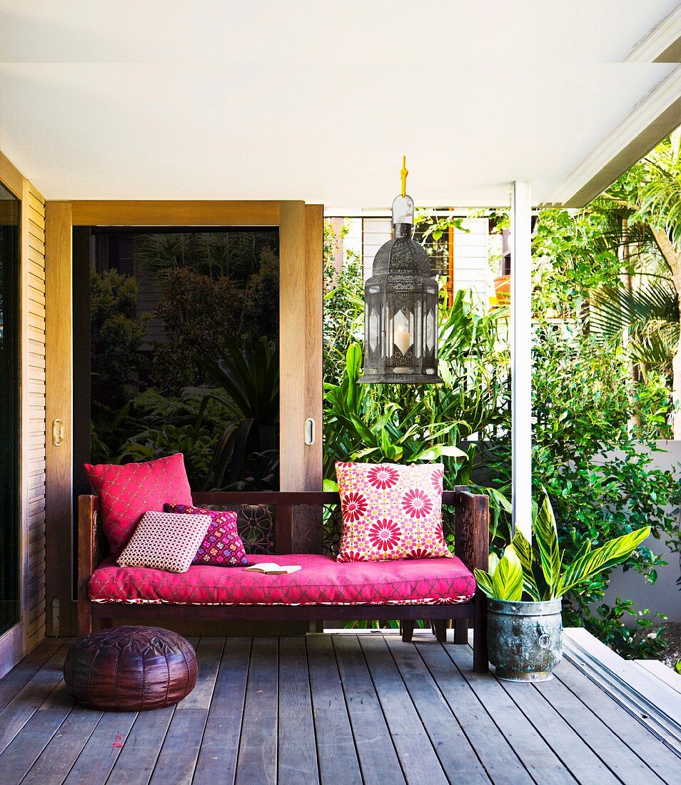 Bench with deep pink seat cushion and scatter cushions on wooden decking; tropical plants in background