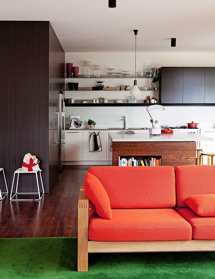 Sofa mit Holzgestell und orangefarbenen Polstern vor der offenen Küche; Einbauten hinter grauer Holzvertäfelung und Kochinsel mit Bücherfach