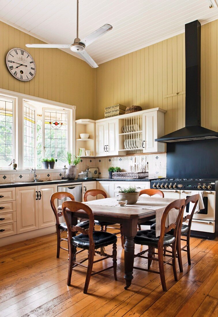 Large, high-ceilinged country-house kitchen with station clock on yellowy beige wall panelling, vintage gas stove with black extractor hood and antique dining set in centre