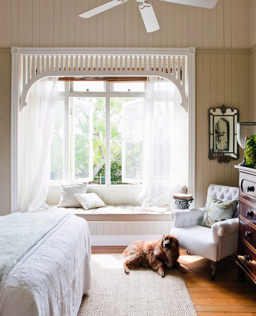 Hund vor einem Polstersessel im Schlafzimmer; romantischer Fenstererker mit Gardinen und integrierter Sitzbank
