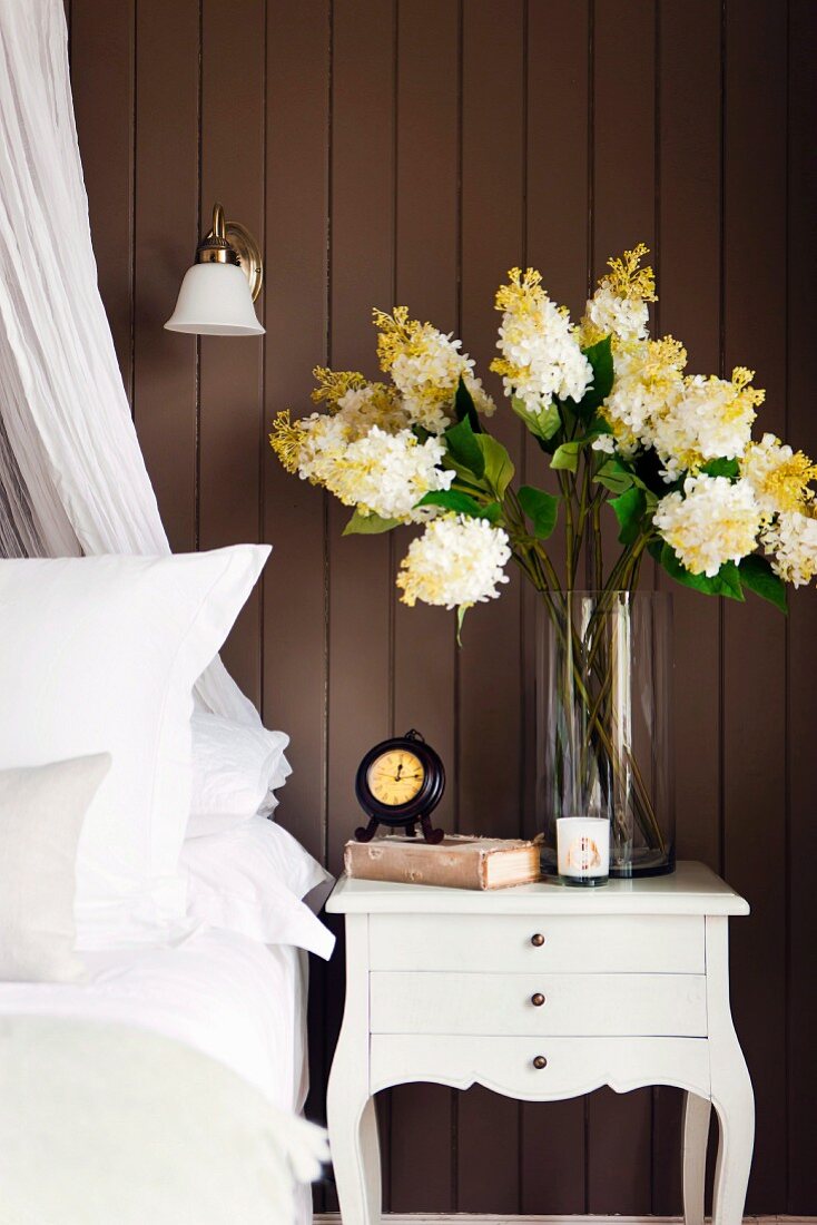 Dunkle Holzvertäfelung hinter blütenweissem Bett mit zierlichem Nachttischchen, Blumenstrauss und antikem Wecker