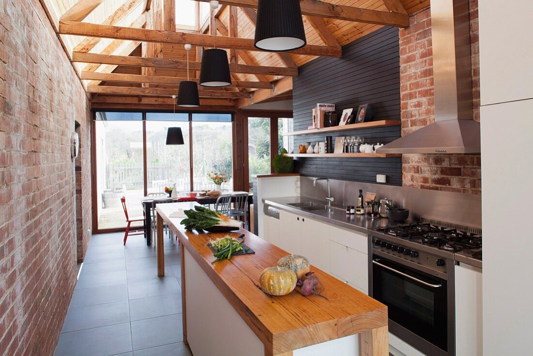 Offene Küche in renoviertem Ziegelhaus mit sichtbarer Dachstuhlkonstruktion aus Holz, daran montierte Hängeleuchten mit schwarzem Lampenschirm