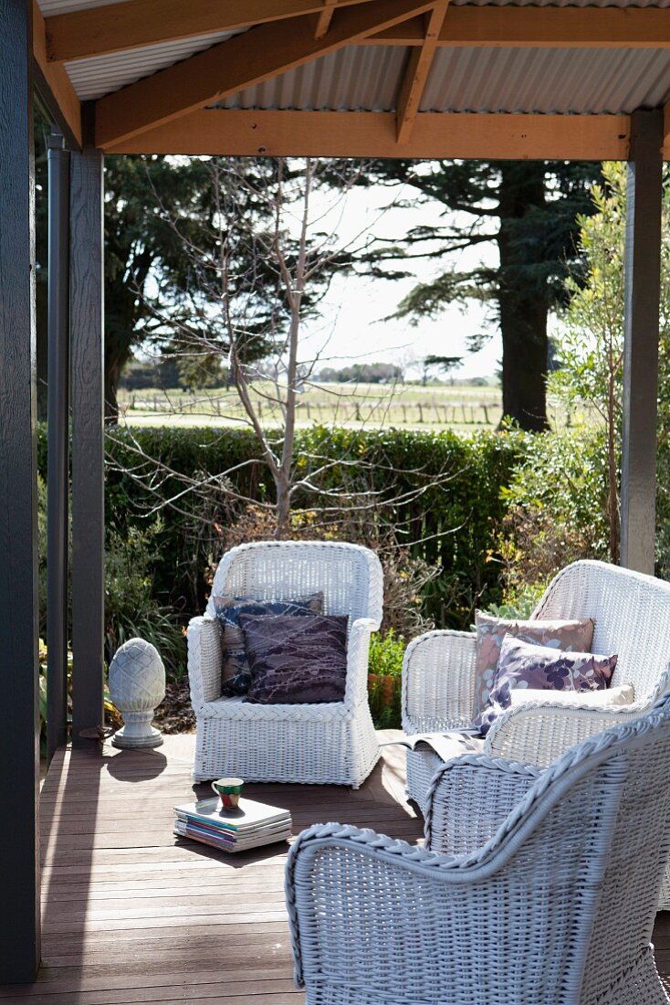 White wicker armchairs on roofed terrace with vintage ambiance; view across sunny garden in rural setting