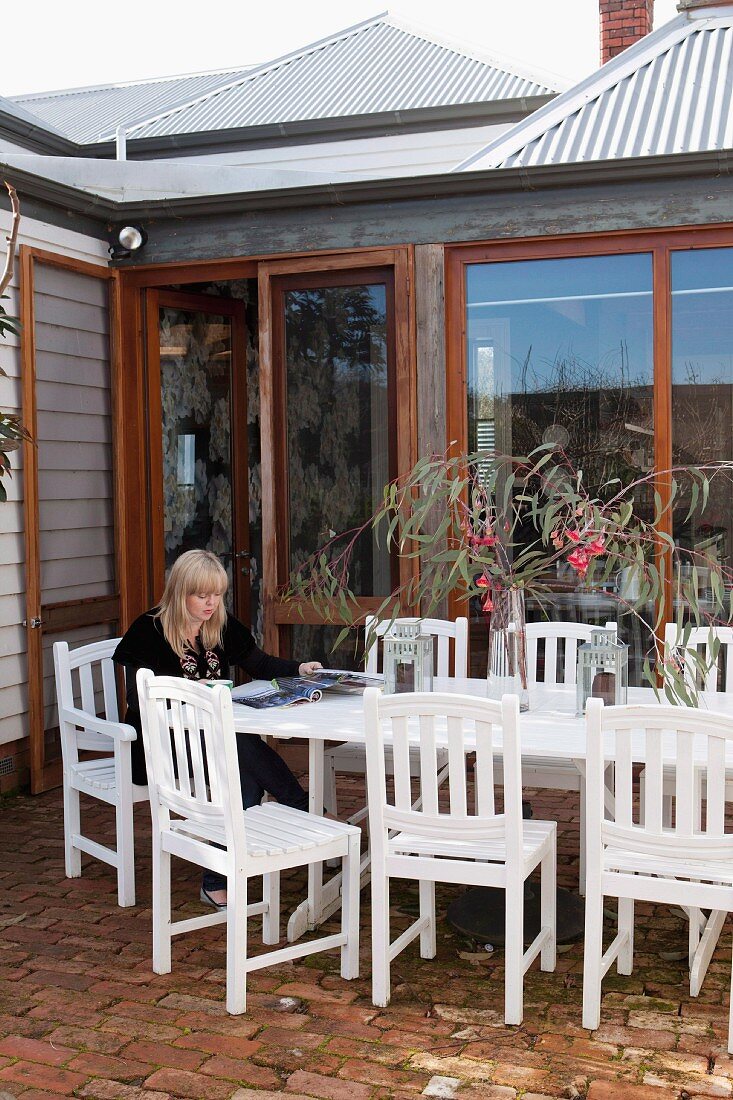 Woman on terrace; white-painted outdoor chairs and table on brick floor outside wooden house with floor-to-ceiling terrace windows