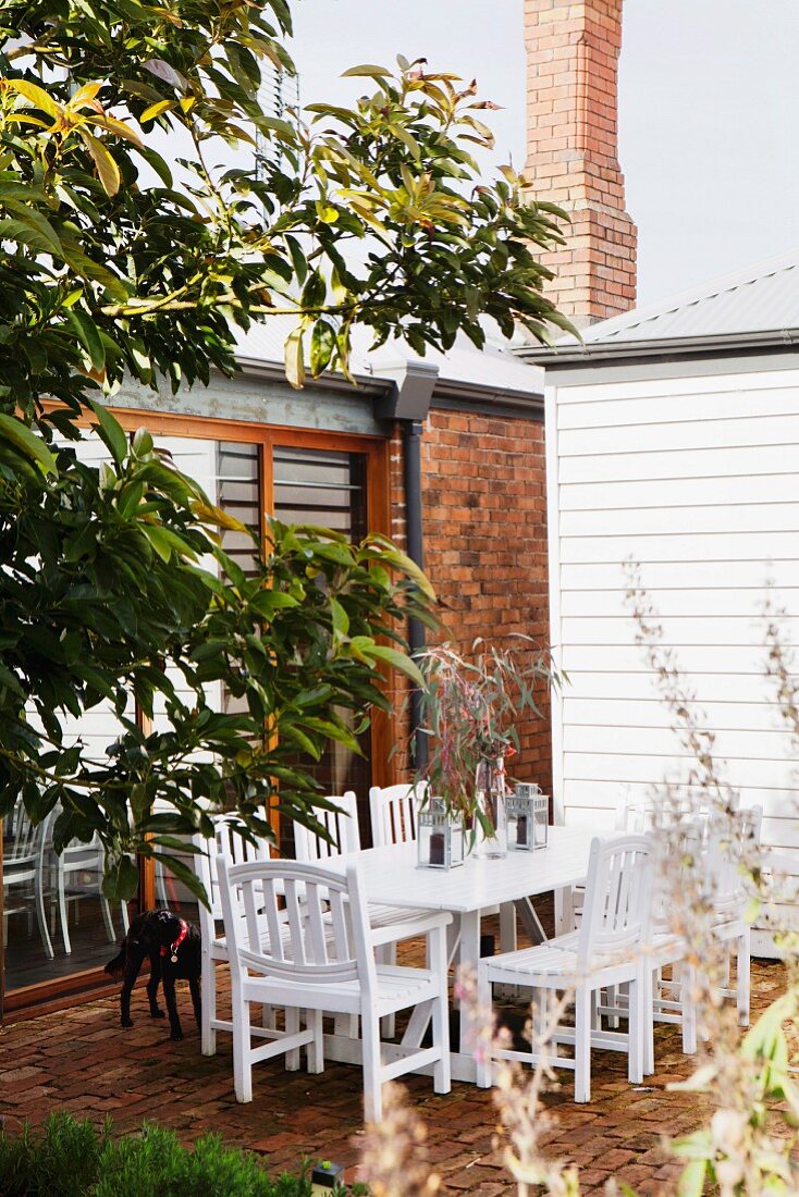 White wooden chairs and table on brick terrace outside house with brick wall, brick chimney and white, wood-clad building to one side