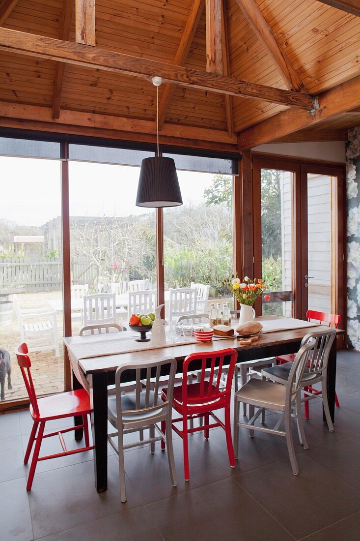 Rote und weiße Stühle an rustikalem Holztisch vor Terrassenfensterfront, sichtbare Holz Deckenkonstruktion