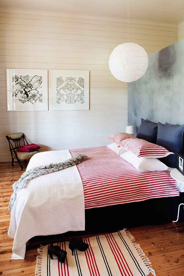 Doppelbett mit rot-weiss gestreifter Bettwäsche vor Raumteiler, im Hintergrund weiße Holzwand in ländlich modernem Schlafzimmer