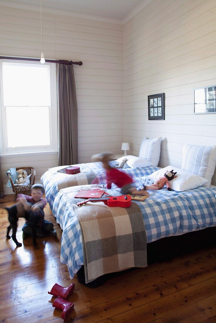Weiß-blau karierte Bettwäsche auf Betten in ländlichem Kinder-Schlafzimmer, weiße Holzverkleidung an Wand; spielende Kinder mit Hund